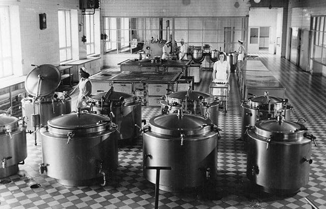 Mustavalkoisessa kuvassa näkyy Seinäjoen piirimielisairaalan keittiötä ja sen työntekijöitä jostain 50-luvulta. tilassa on useita puuhelloja sekä isoja ruuanvalmistukseen tarvittavia patoja.